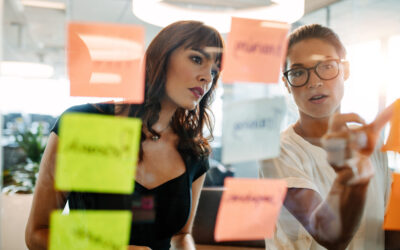 Creative professionals brainstorming on new business ideas Asian businesswoman showing her coworker an idea posted on a sticky note wall. Creative professionals brainstorming on new business ideas.